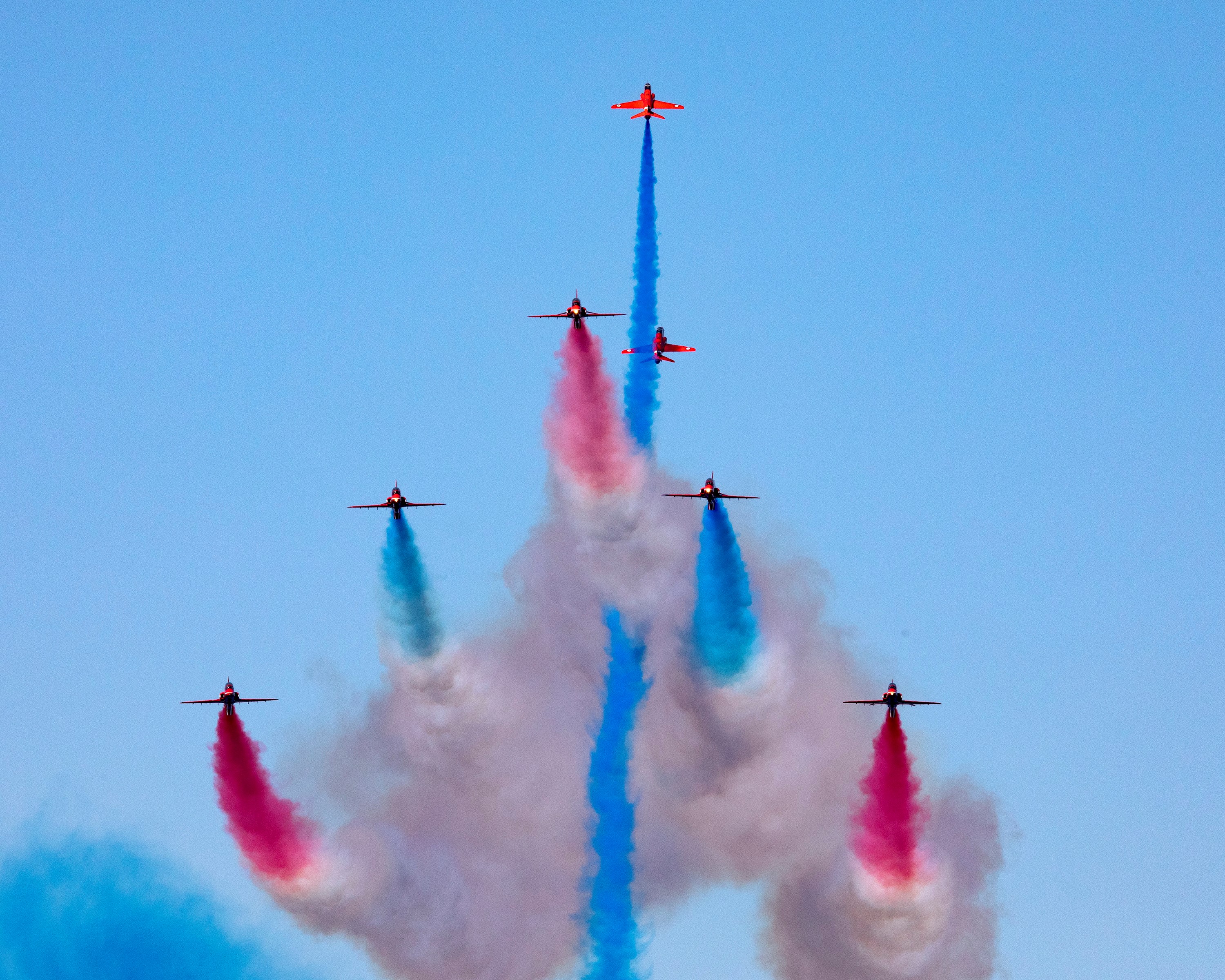 Image shows the Red Arrows performing in formation, with red, blue and white smoke trails. 